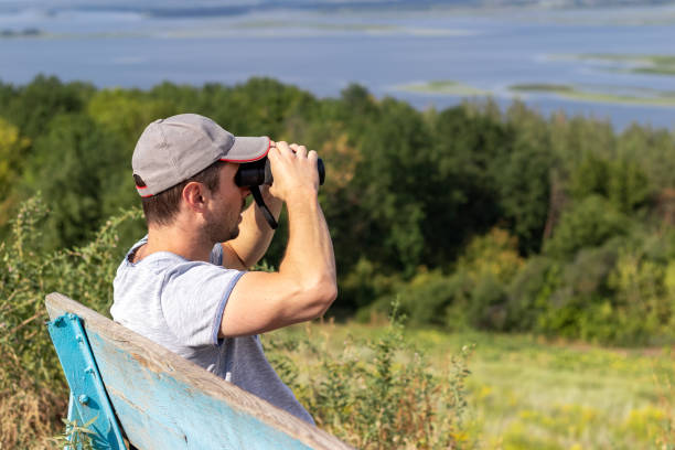 istock A man looks through binoculars at a scenic view of a wide river while sitting on an old wooden bench on a hill. Side view 1268441729