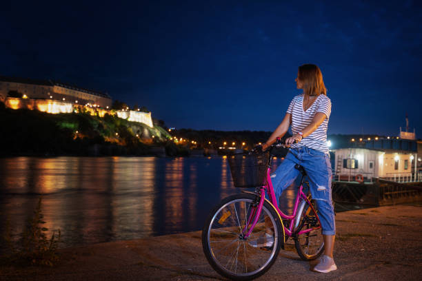 Young woman with a bike on the Novi Sad embankment overlooking the Petrovaradin fortress, night cityscape travel to Serbia, Balkan countries Young woman with a bike on the Novi Sad embankment overlooking the Petrovaradin fortress, night shot, travel to Serbia, Balkan countries Petrovaradin stock pictures, royalty-free photos & images