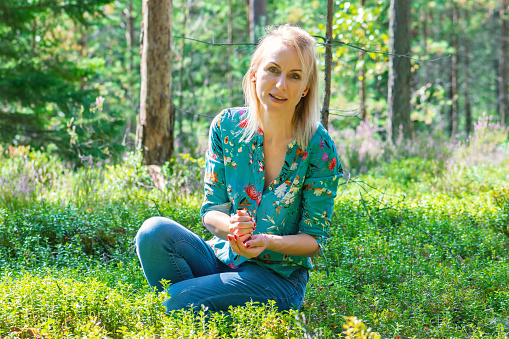 Blonde woman with colorful blouse collecting cranberries in dense green forest and illuminated by summer sun beams