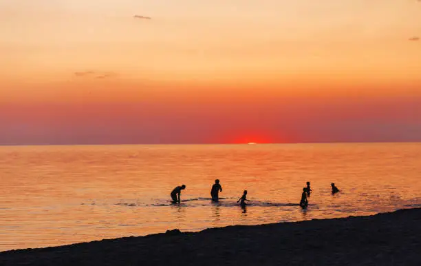 June 26, 2020 - Palanga, Lithuania: amazing orange red sunset at the sea with several silhouettes of people taking last swim, enjoying evening with only the tip of the sun visible in the horizon.
