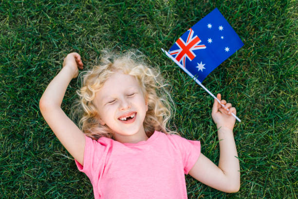 Adorable cute happy Caucasian girl holding Australian flag. Smiling laughing child lying on grass in park holding Australia flag. Kid citizen celebrating Australia Day holiday in January outdoor. adorable lindo feliz chica caucásica sosteniendo la bandera australiana. niño riendo sonriendo tumbado en el césped en el parque sosteniendo la bandera de australia. ciudadano menor celebrando las vacaciones del día de australia en enero al aire libre.