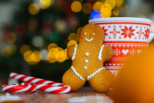 Christmas postcard with gingerbread man red mug with christmas ornament on light bokeh from Xmas tree and red striped holidays candies. Xmas candies and cup with marshmallow on wood background