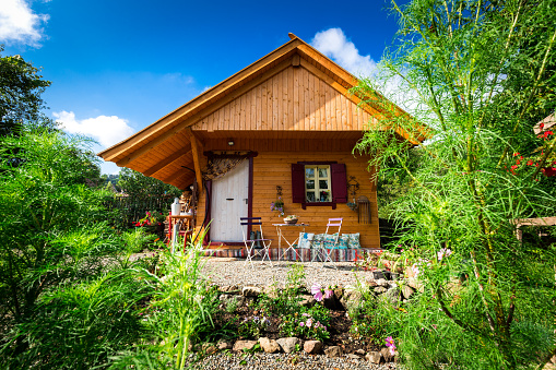 Wide angle color image depicting a small, cosy wooden cabin in a garden. The cabin has a table and chairs in front of it for relaxation. Room for copy space.