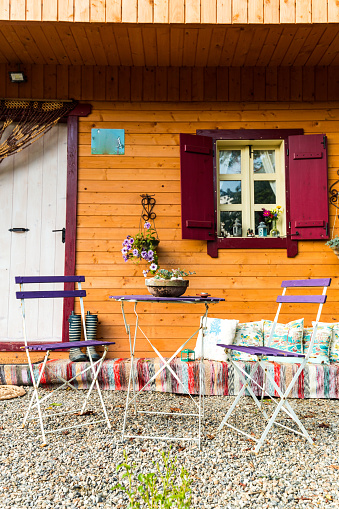 Wide angle color image depicting a small, cosy wooden cabin in a garden. The cabin has a table and chairs in front of it for relaxation. Room for copy space.