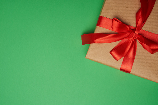 rectangular box wrapped in brown paper and tied with a silk red ribbon with a bow, gift on a green background, top view, copy space