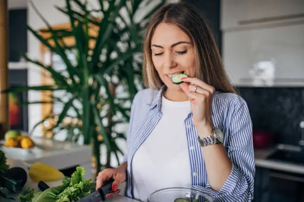 One woman, beautiful young woman in kitchen is preparing a vegan salad in casual clothes, she is tasting a cucumber.