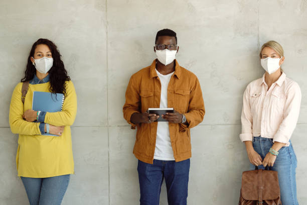 Multi-ethnic group of college students with protective face masks against the wall. Smiling group of university students wearing protective face masks while standing by the wall in a hallway. kn95 face mask photos stock pictures, royalty-free photos & images