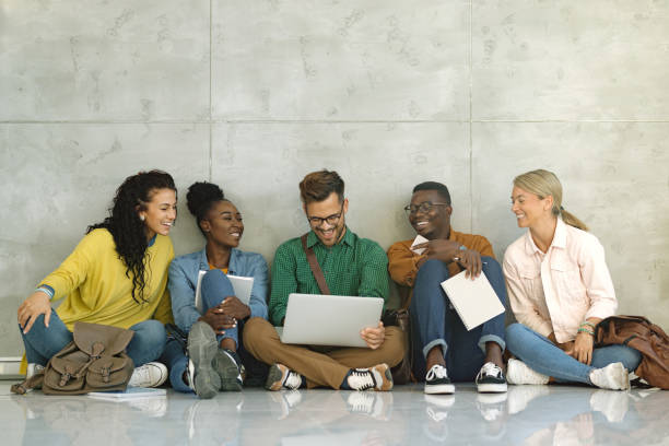 Happy university students using laptop while sitting in a hallway. Group of happy students surfing the net on laptop while sitting on the floor at university hallway. Copy space. sitting on floor stock pictures, royalty-free photos & images