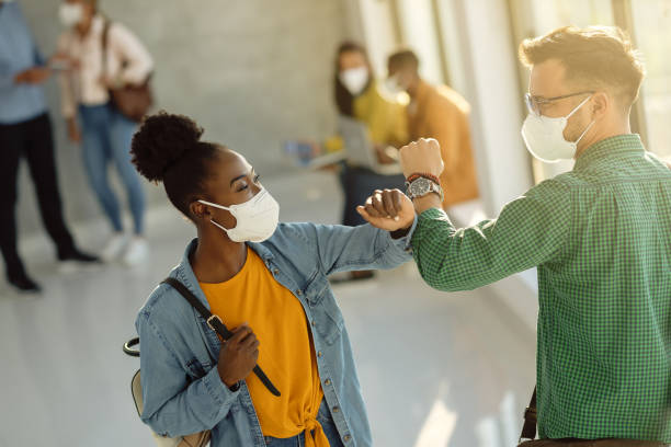 Happy university students greeting with elbow while wearing protective face masks. Happy college friends with protective face masks elbow bumping in a hallway. college students studying together stock pictures, royalty-free photos & images