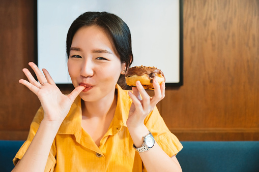 Asian woman is finger-licking good while having a chocolate glazed homemade donut topping with crush hazelnut in a modern cafe. Enjoyment female lifestyle.