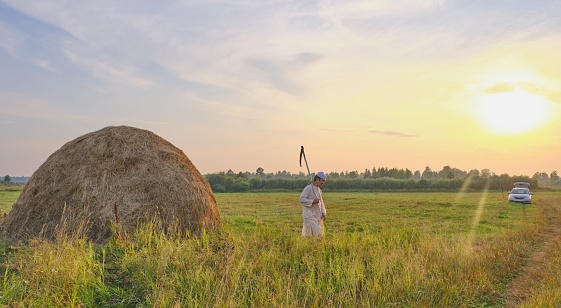 An senior Muslim man in a skullcap and traditional clothes leaves with a hand scythe from the hay field at sunset. Country life concept