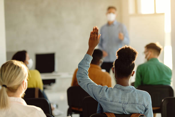 Rear view of African American student raising hand during a class in lecture hall. Back view of black female student raising arm to ask a question during a lecture in the classroom. continuing education stock pictures, royalty-free photos & images