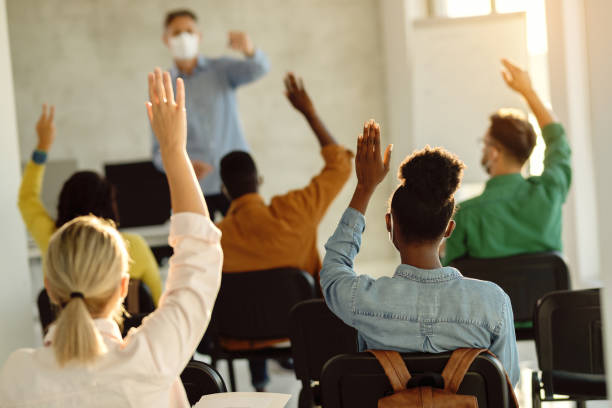 Back view of group of students raising their arms during a class at lecture hall. Rear view of college students raising hands to answer teacher's question during a lecture. adult student stock pictures, royalty-free photos & images
