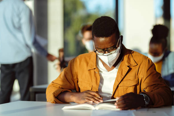 African American college student with face mask reading a book in the classroom. Black student wearing protective face mask while studying in lecture hall during coronavirus epidemic. kn95 face mask photos stock pictures, royalty-free photos & images