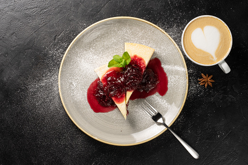 A cup of cappuccino and two slices of cheesecake on a round plate drizzled with strawberry jam and garnished with icing sugar and mint leaves.