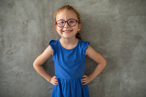 Redhead cross-eyed girl standing in front of gray wall at home, looking at camera and smiling, she's wearing blue dress