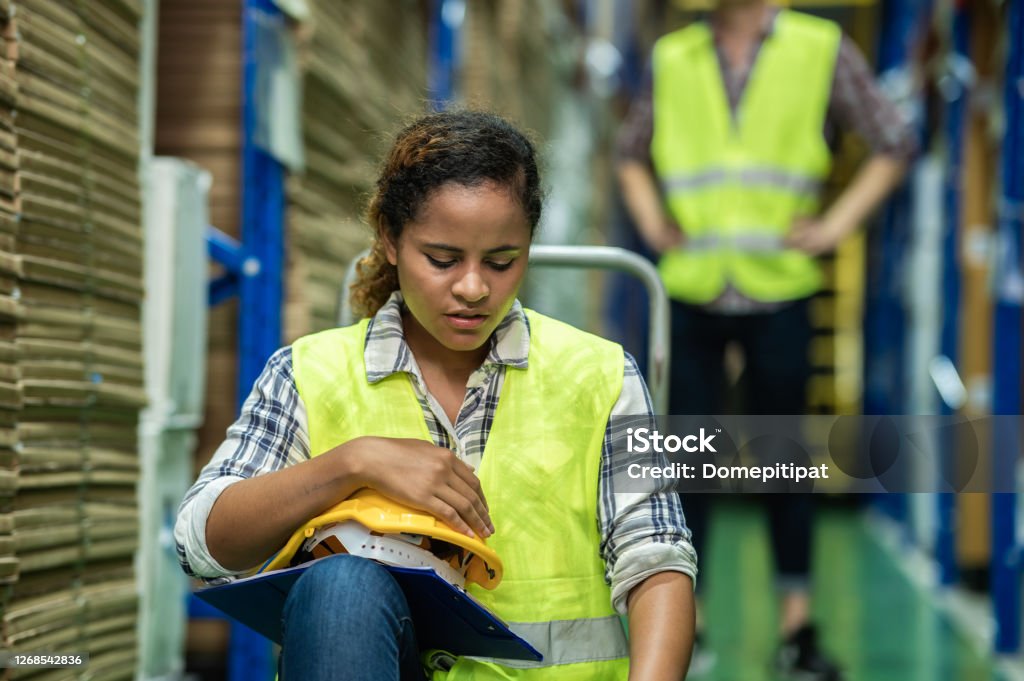 Young African female warehouse worker staff feeling sad and upset while sitting on the floor of the storehouse due to been fired from job cause by company bankruptcy from coronavirus pandemic. Being Fired Stock Photo