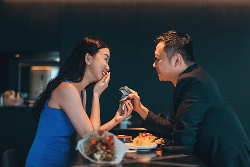 Asian man proposing over a meal during Covid pandemic. Social distance markers are still visible on restaurant tables and in public spaces