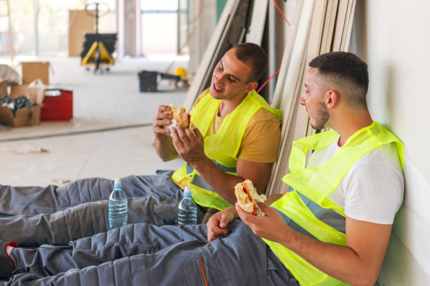 Taking Break from Work Two workers wearing safety helmets take a break from work and enjoy lunch while sitting inside, an unfinished building in the background construction lunch break stock pictures, royalty-free photos & images