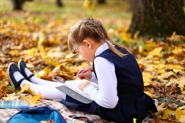 istock Back to elementary school. Girl in dress does homework with pen in hand.Kid study on plaid blanket.Picnic in park on maple yellow leaves.Blue backpack,notebooks,textbooks lie on ground.Autumn concept 1268568728