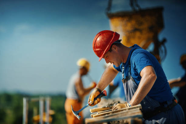 Closeup side view of a construction worker making a wooden mold before pouring concrete on the last floor of an apartment building. Couple of his colleagues in background and guiding concrete pouring machine.