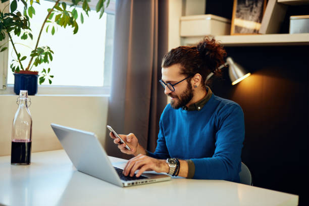 istock Young attractive hipster sitting in his home office and using smart phone during corona virus. 1268571072