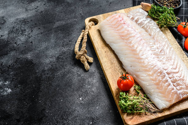 istock Raw cod fillet with and herbs on rustic wooden cutting board.  Black background. Top view. Copy space 1268573281
