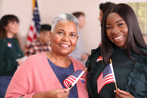 African descent mother, daughter or friends vote in the USA election.  They stand together holding flags and wearing \