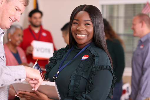 African descent, young adult woman volunteer works at polling place in the USA election. She assists mature man with his voter registration.
