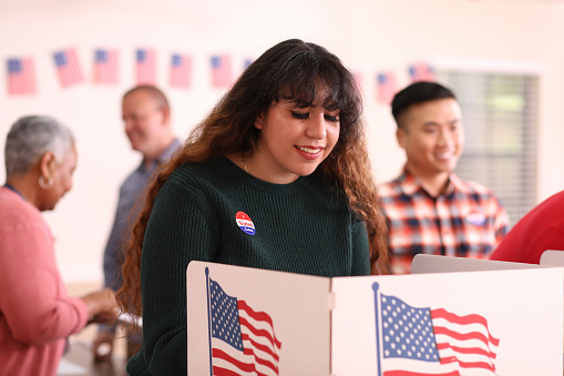 Latin descent, young adult woman votes in the USA election.  She stands at voting booth in polling station.   Other voters and election day registration seen in background.