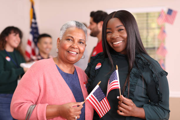 Friends or mother, daughter are all smiles as they vote in USA election. African descent mother, daughter or friends vote in the USA election.  They stand together holding flags and wearing "I Voted" stickers inside their local polling station.   Other voters and election day registration seen in background. citizenship stock pictures, royalty-free photos & images