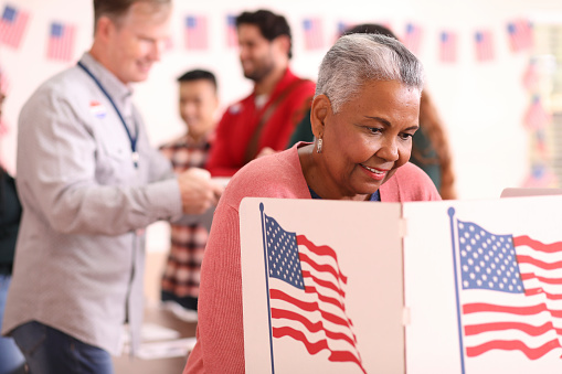 African descent, senior adult woman votes in the USA election.  She stands at voting booth in polling station.   Other voters and election day registration seen in background.