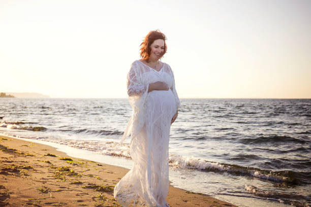 A pregnant woman walking on a beach in a beautiful lace dress with the setting sun and the blue ocean behind her.