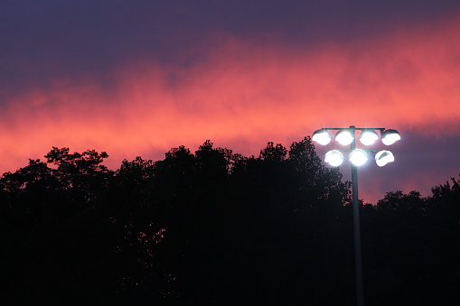 The stadium lights shine a brilliant white against a backdrop of purples and oranges during sunset, atop a tree line silhouette. Waiting for football