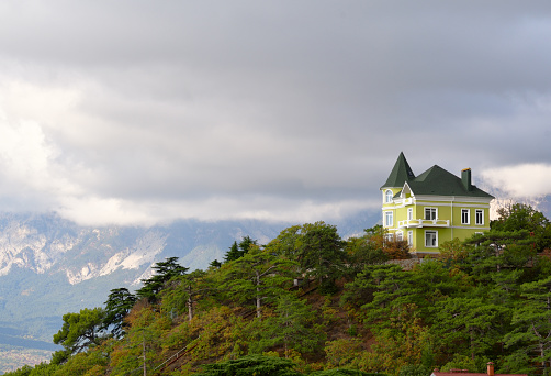 A house with a turret on top of a hill among green trees. Mountains drowned in clouds in the distance. Russia, Crimea, 2019