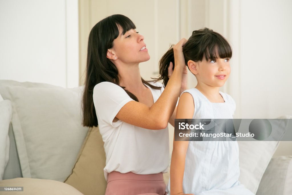 Young Caucasian mother holding hairs of her daughter Young Caucasian mother holding hairs of her daughter. Pretty brunette mom sitting on sofa and doing ponytail hairstyle for little cute girl. Family time and parenthood concept Girls Stock Photo