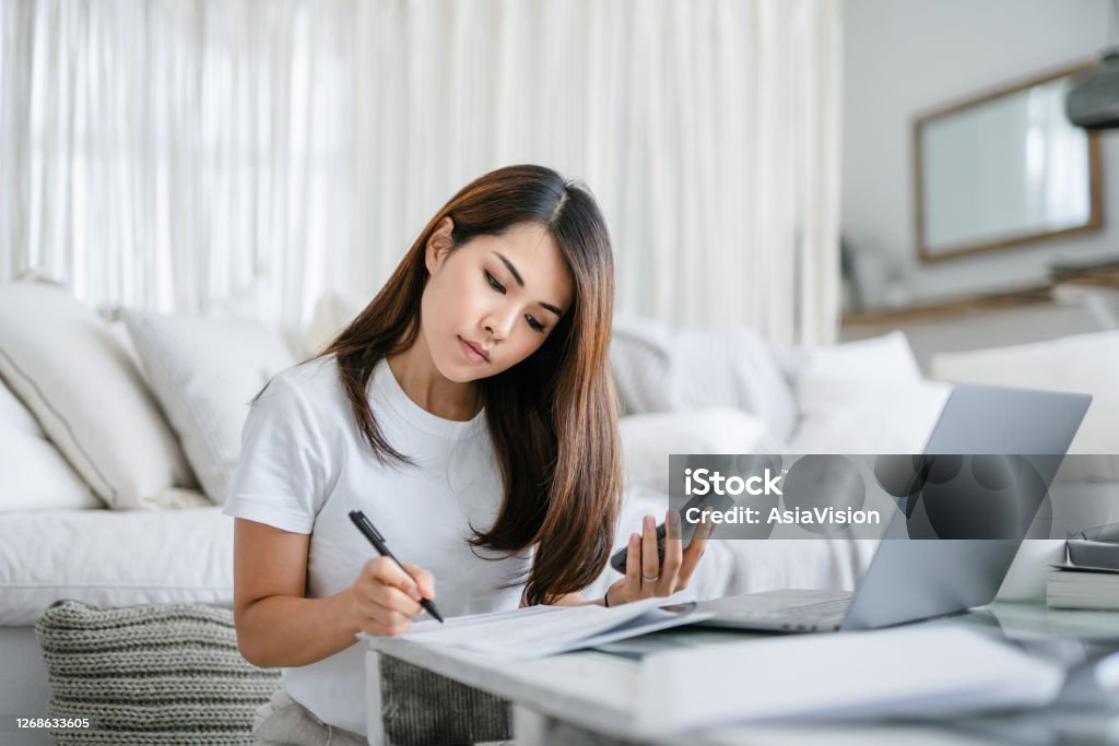 Young Asian woman sitting on the floor by the sofa in the living room working from home using laptop computer and handling paperworks Finance Stock Photo