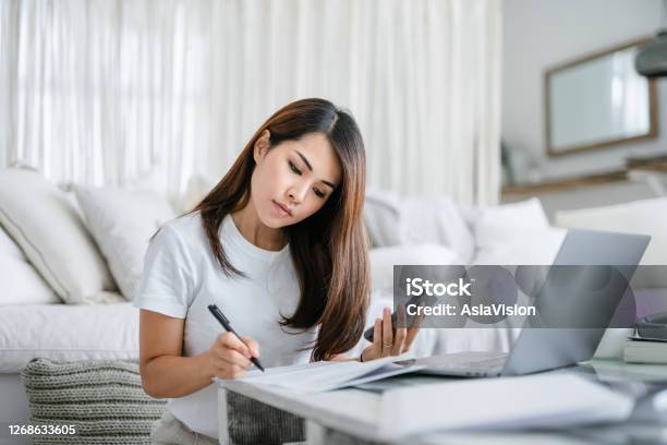 Young Asian Woman Sitting On The Floor By The Sofa In The Living Room Working From Home Using Laptop Computer And Handling Paperworks Stock Photo - Download Image Now
