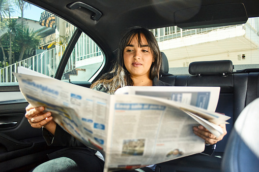 Young mixed race businesswoman reading the newspaper while wearing white blouse and black blazer jacket in the morning sitting in the back seat of a taxi with newspaper