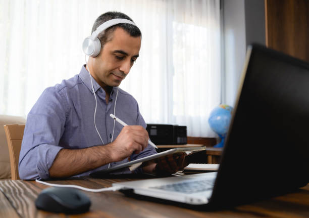 istock Man Is Working Online In Home Office Using His Laptop Computer, Joining Webinar And Taking Notes On Digital Tablet 1268651650