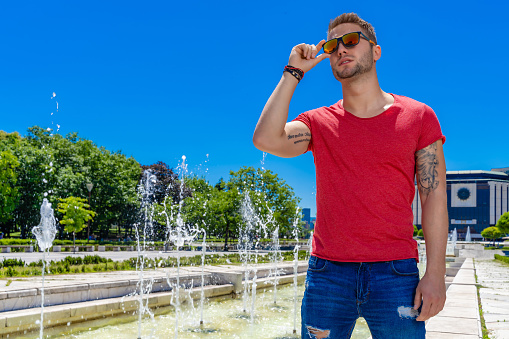 Young adult adjusting his sunglasses in front fountain and National Palace of Culture - NDK, Sofia, Bulgaria (Bulgarian: Национален дворец на културата - НДК ).  The scene is situated outdoors in front NDK, Sofia,  Bulgaria. Photo is taken with SONY A7III camera