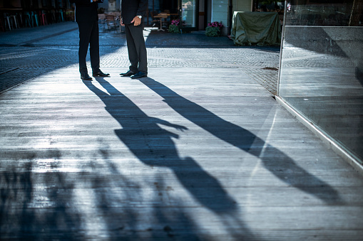 Partial view of male corporate professionals casting long shadows as they stand and talk outdoors in late afternoon.