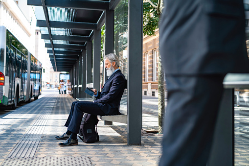 Low angle personal perspective of bearded corporate professional in mid 50s sitting on bench at bus stop and checking smart phone while he waits.