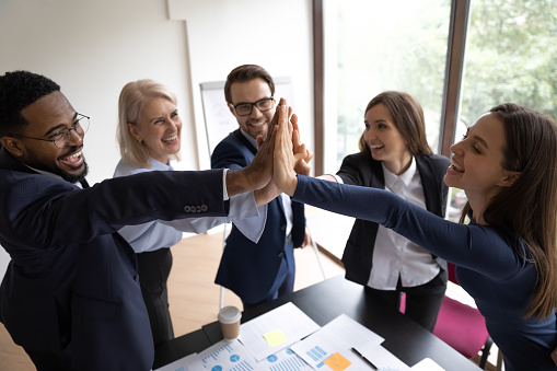 Happy diverse employees coworkers or business partners joining hands in high five gesture celebrating success or victory, feeling motivated, proud and satisfied of teamwork and common achievements