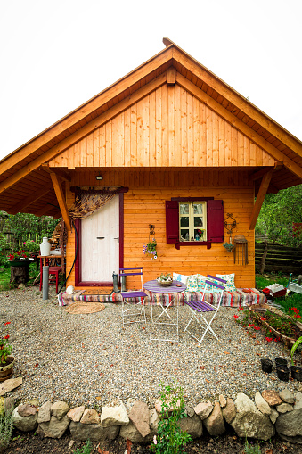 Wide angle color image depicting a small, cosy wooden cabin in a garden. The cabin has a table and chairs in front of it for relaxation. Room for copy space.