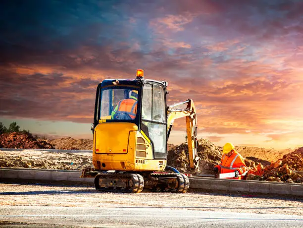 Photo of Mini Excavator and worker at a construction site against the setting sun