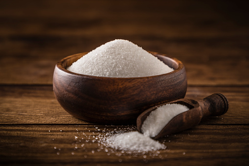 Front view of a wooden bowl full of white sugar beside a serving scoop against a rustic wooden table. Predominant color is brown. Studio shot taken with Canon EOS 6D Mark II and Canon EF 24-105 mm f/4L