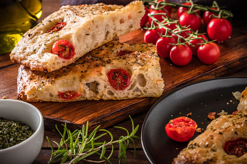 Front view of two sliced homemade Italian focaccia. The focaccia is on a wooden cutting board and is surrounded by some ingredients like an olive oil bottle, cherry tomatoes, a bowl full of dried oregano and some fresh rosemary leaves. Predominant colors are brown and red. Studio shot taken with Canon EOS 6D Mark II and Canon EF 24-105 mm f/4L