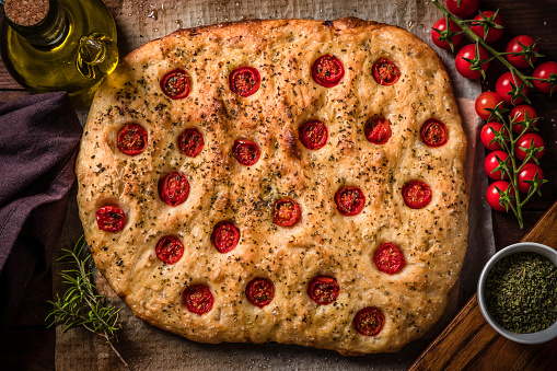 Top view of a homemade Italian focaccia. The focaccia is at the center of the image and is surrounded by some ingredients like an olive oil bottle, cherry tomatoes, a bowl full of dried oregano and some fresh rosemary leaves. Predominant colors are brown and red. Studio shot taken with Canon EOS 6D Mark II and Canon EF 24-105 mm f/4L