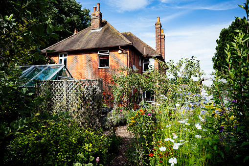 Garden view of footpath leading through summertime flowers, fruit trees, and greenhouse to two-story brick home on summer day.